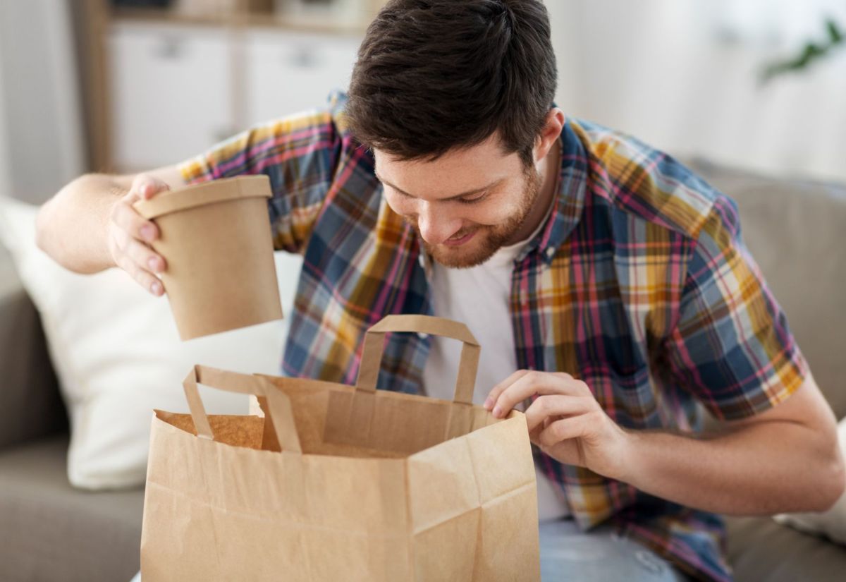 Hombre sentado en el sofá abriendo una bolsa de cartón con táper de comida a domicilio