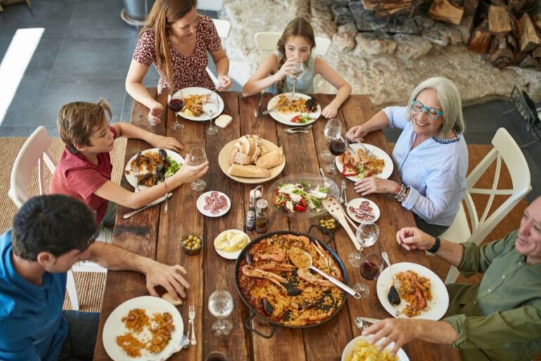 Comida familiar con mesa llena de comida y paella en el centro