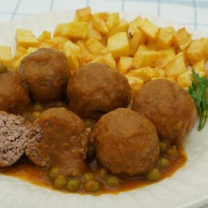 Plato de albóndigas acompañadas de patatas fritas cortadas en cuadraditos, guisantes y una ramita de perejil