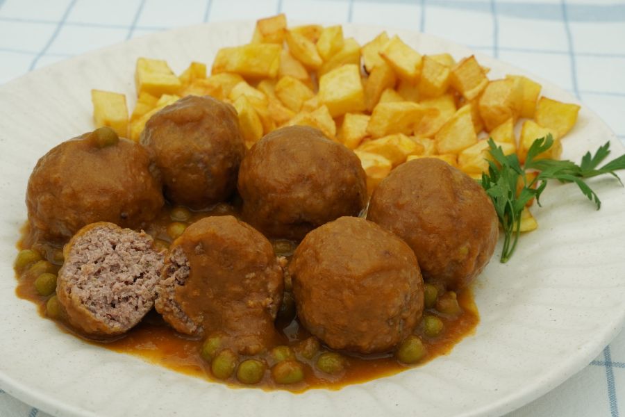 Plato de albóndigas acompañadas de patatas fritas cortadas en cuadraditos, guisantes y una ramita de perejil
