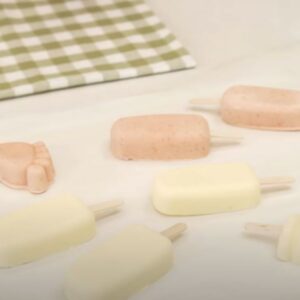 Helados de diferentes sabores y formas distribuidos sobre una mesa blanca con un paño de cocina de cuadros verdes y blancos al fondo