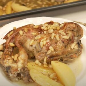 Plato con pierna de cordero asada acompañada de salsa, patatas y piñones